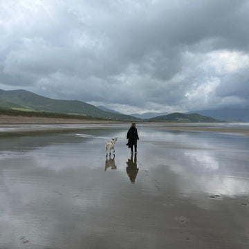 West Coast Crafts Nicole walking her dog Bowie on Brandon Bay beach in Kerry with mountains in the background under a cloudy sky