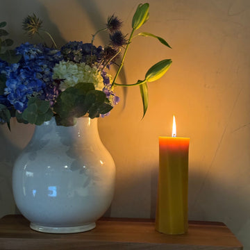White vase with flowers and a lit yellow West Coast Crafts beeswax candle on a wooden surface.